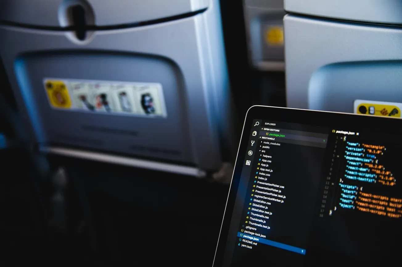 Open laptop displaying colorful code sits on an airplane tray table. The plane's seatbacks are visible, creating a focused yet casual work-in-transit vibe.