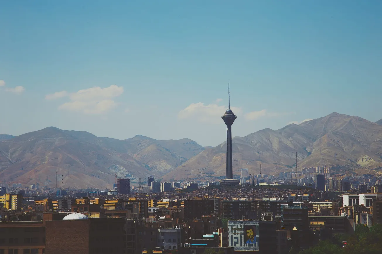 Cityscape of Tehran with the Milad Tower prominently rising against the backdrop of the Alborz Mountains under a clear blue sky.