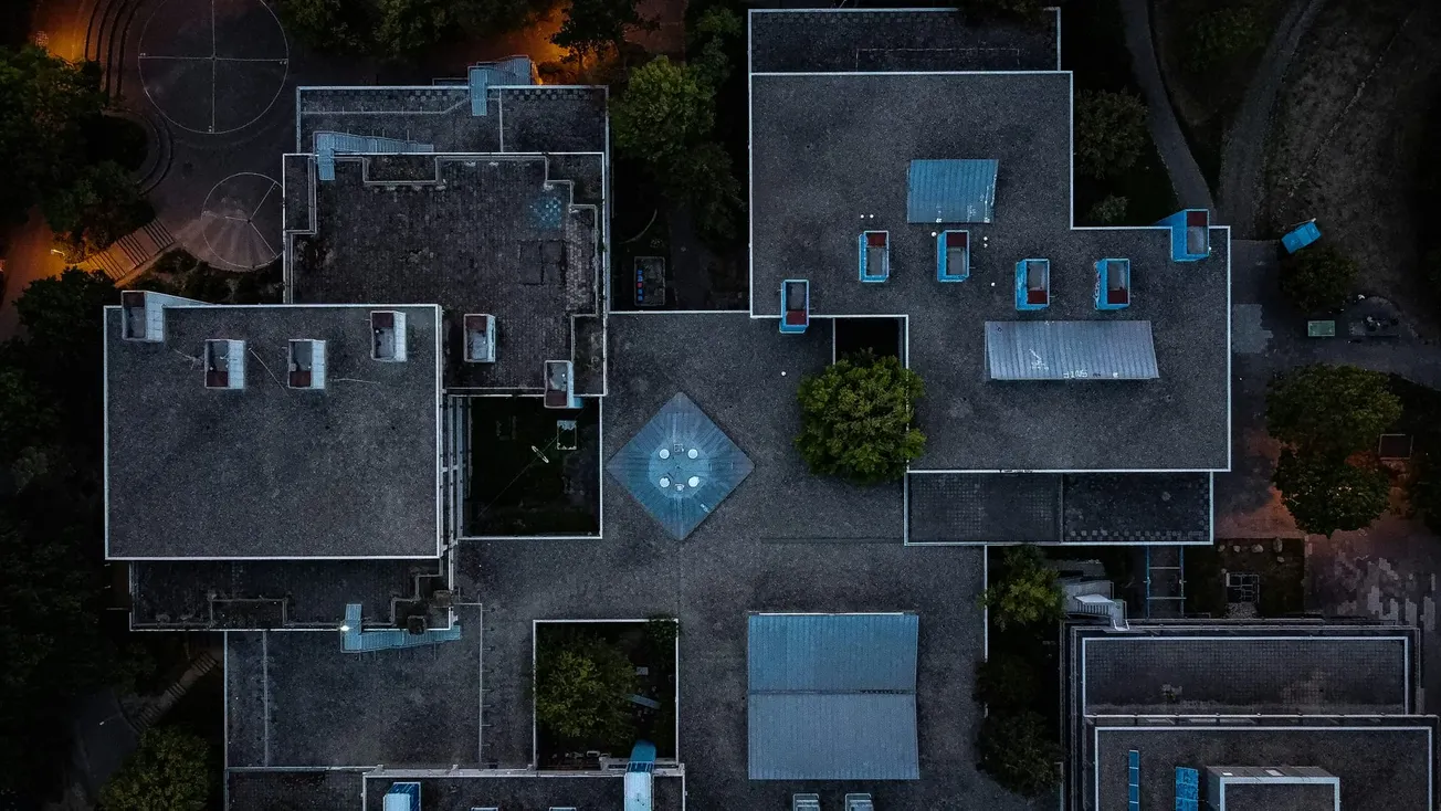 Aerial view of an urban building complex at night, featuring interconnected flat roofs and dim streetlights, surrounded by trees. The scene is tranquil and geometric.