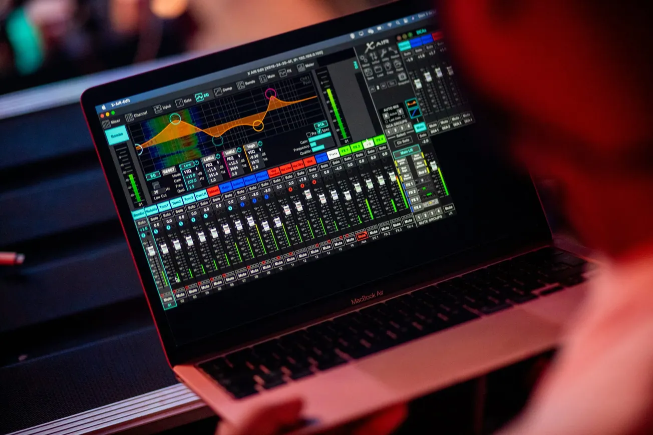 A person operates a laptop displaying colorful audio mixing software with various equalizer and volume controls, conveying a technical, professional tone.