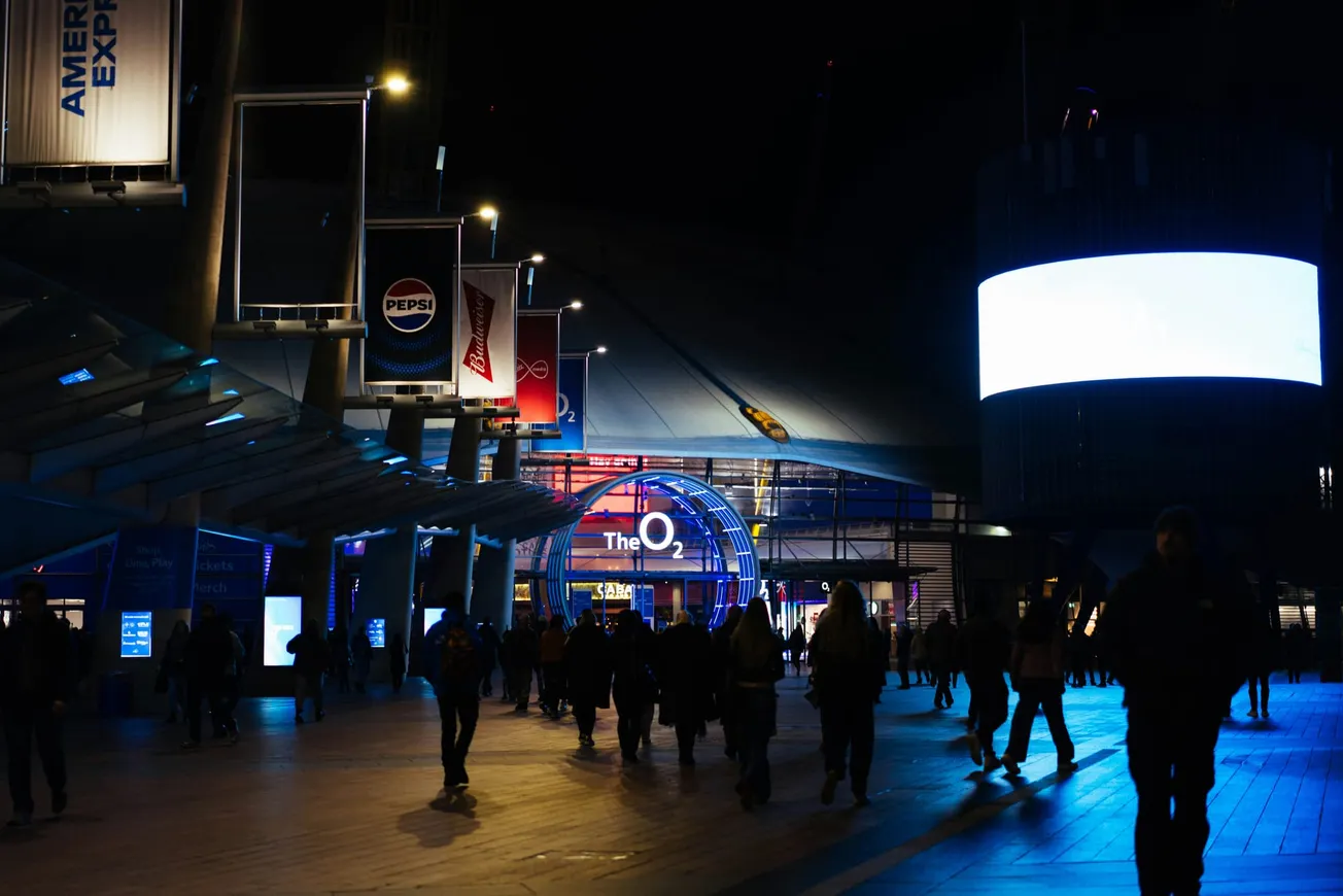 Crowd walking towards the illuminated entrance of The O2 arena at night, with vibrant blue and red lights, advertising banners overhead.
