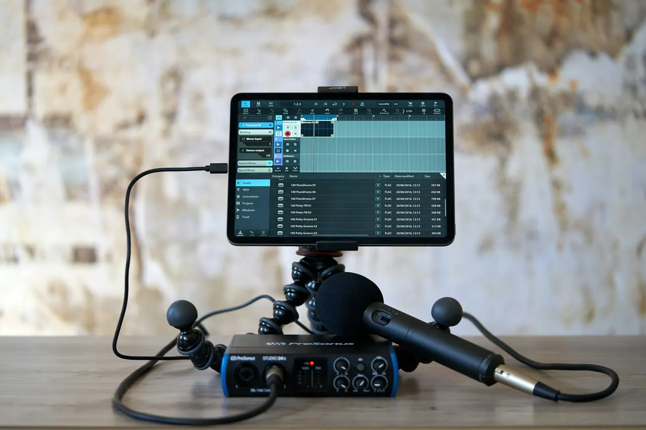 A tablet on a flexible tripod displays audio editing software. A microphone and audio interface are in the foreground on a wooden table. The backdrop is a textured, rustic wall. 