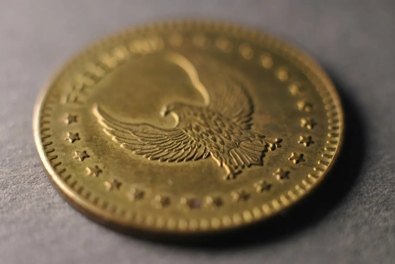 Close-up of a golden coin featuring an embossed eagle with spread wings surrounded by stars along the edge, resting on a flat surface.