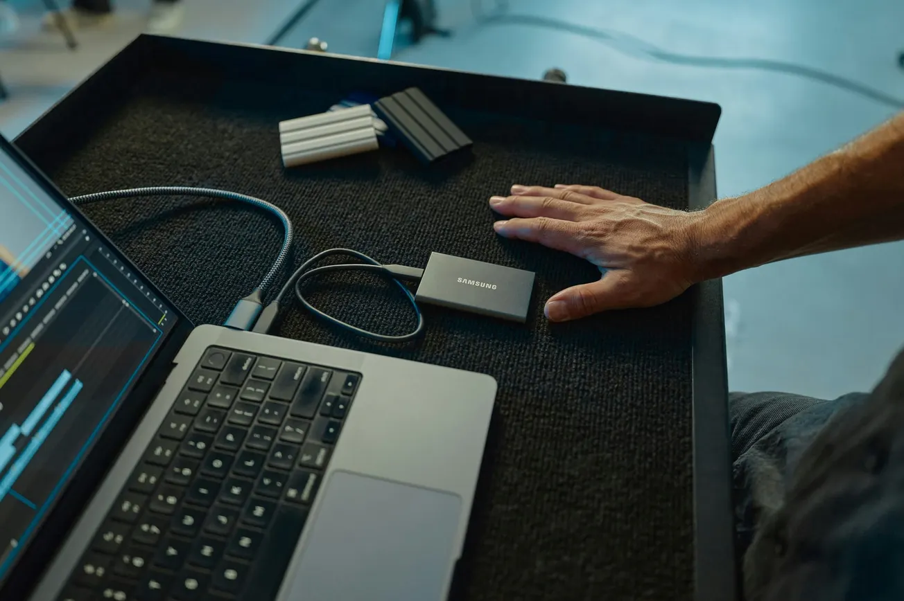 A person’s hand rests beside an external hard drive connected to a laptop displaying video editing software. Nearby are three other hard drives.