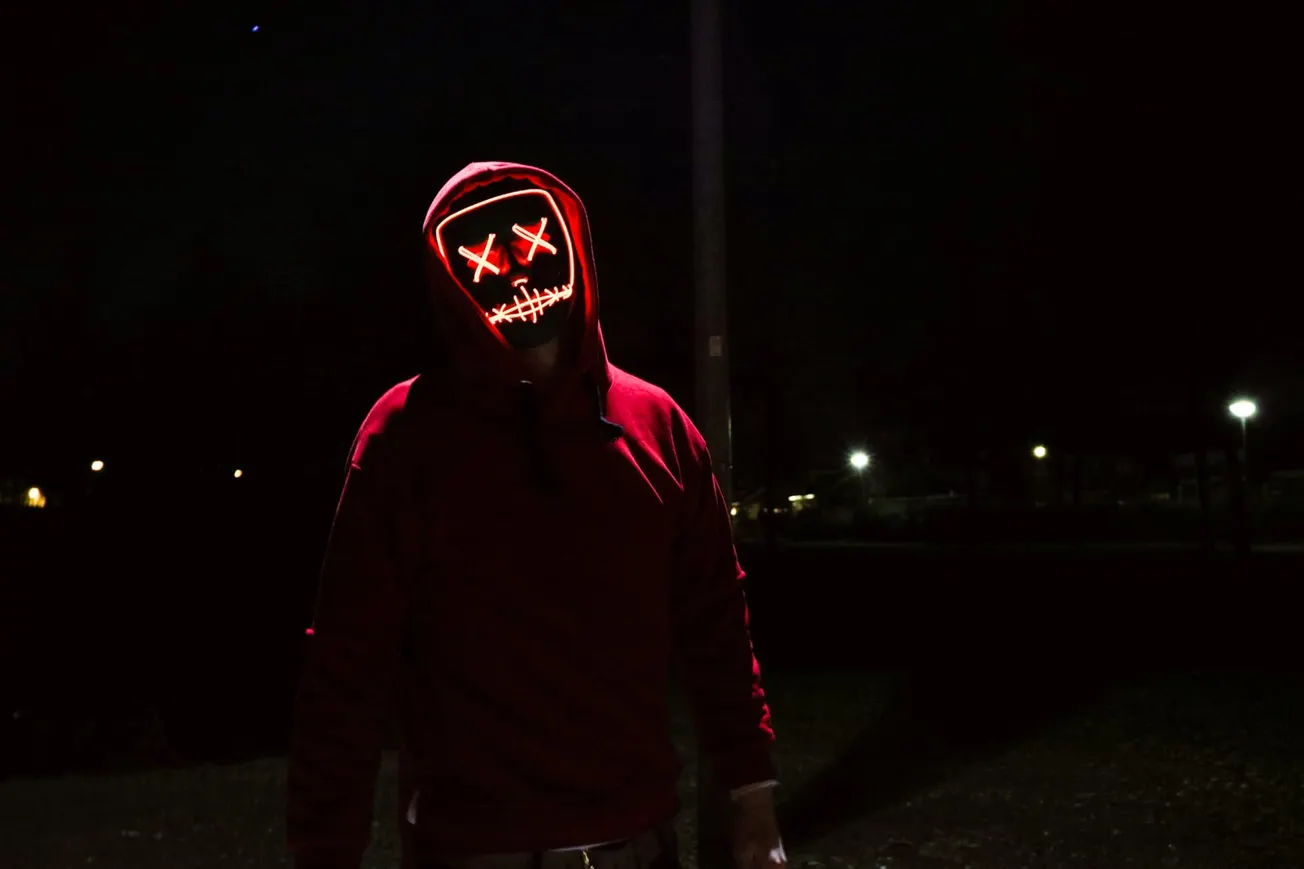 A person in a red hoodie stands at night, wearing a glowing mask with red X eyes and a stitched smile, exuding a mysterious, eerie vibe.
