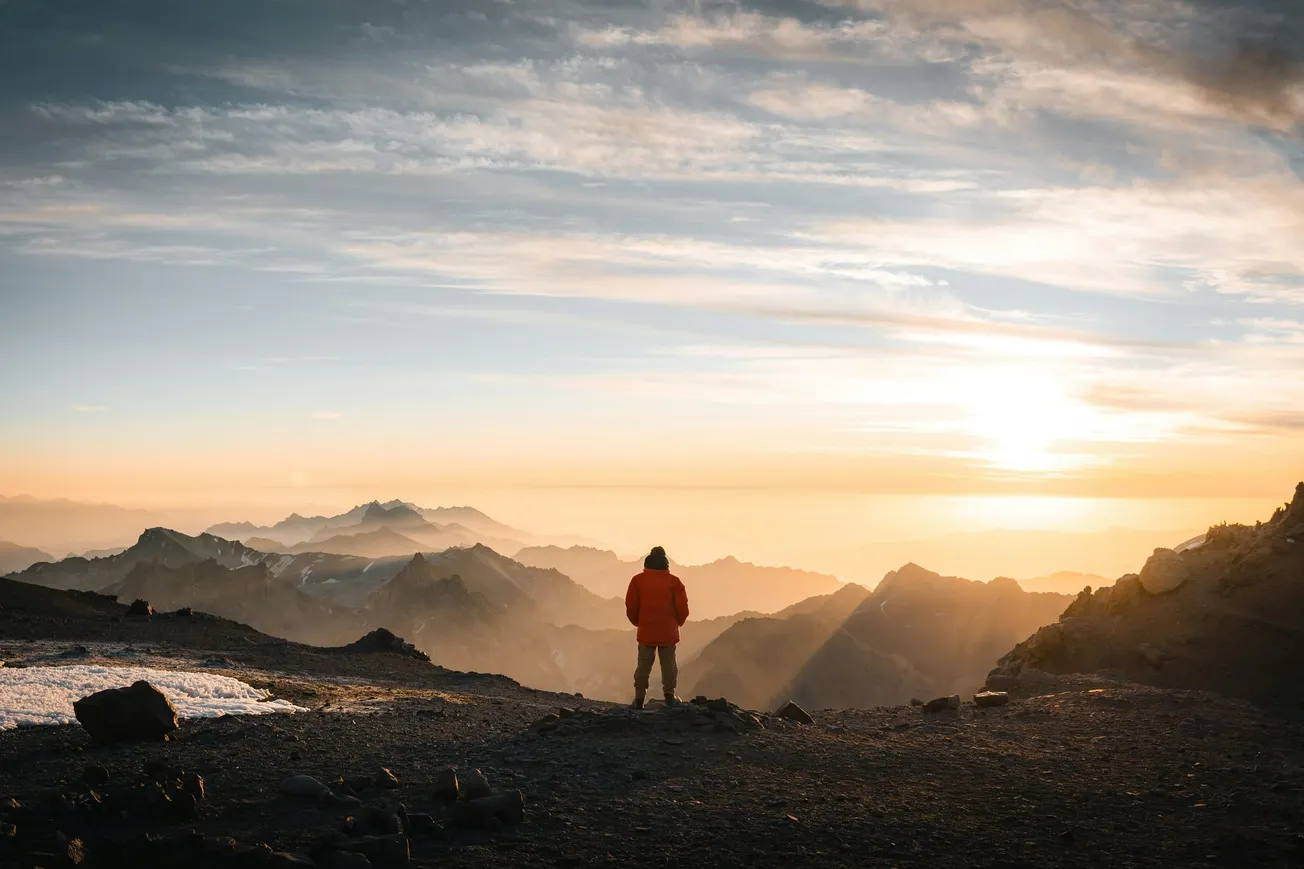 A person in a red jacket stands on a mountain peak, overlooking a vast range during sunrise. The sky is filled with soft clouds and golden light.