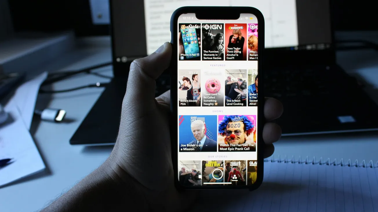 A hand holds a smartphone displaying a colorful news app with various headlines. A laptop and notebook are visible in the blurred background.