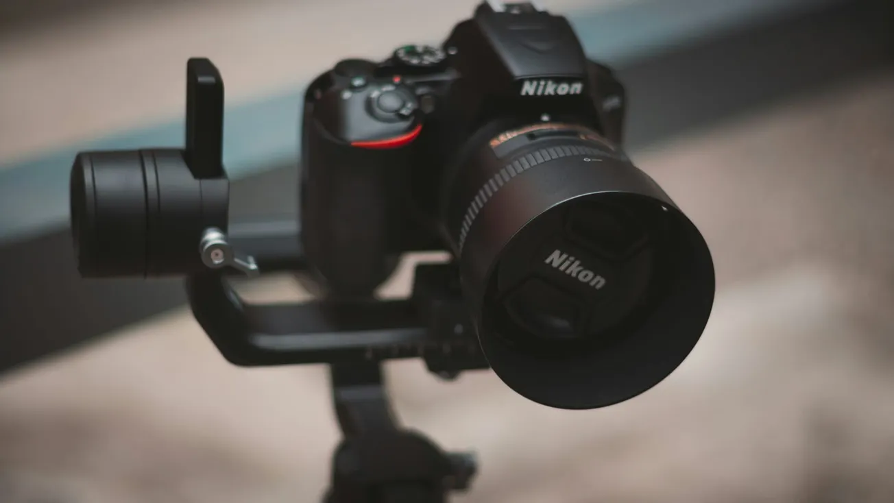 Close-up of a Nikon DSLR camera mounted on a stabilizer, with a prominent lens and a blurred background, conveying a professional photography setup.