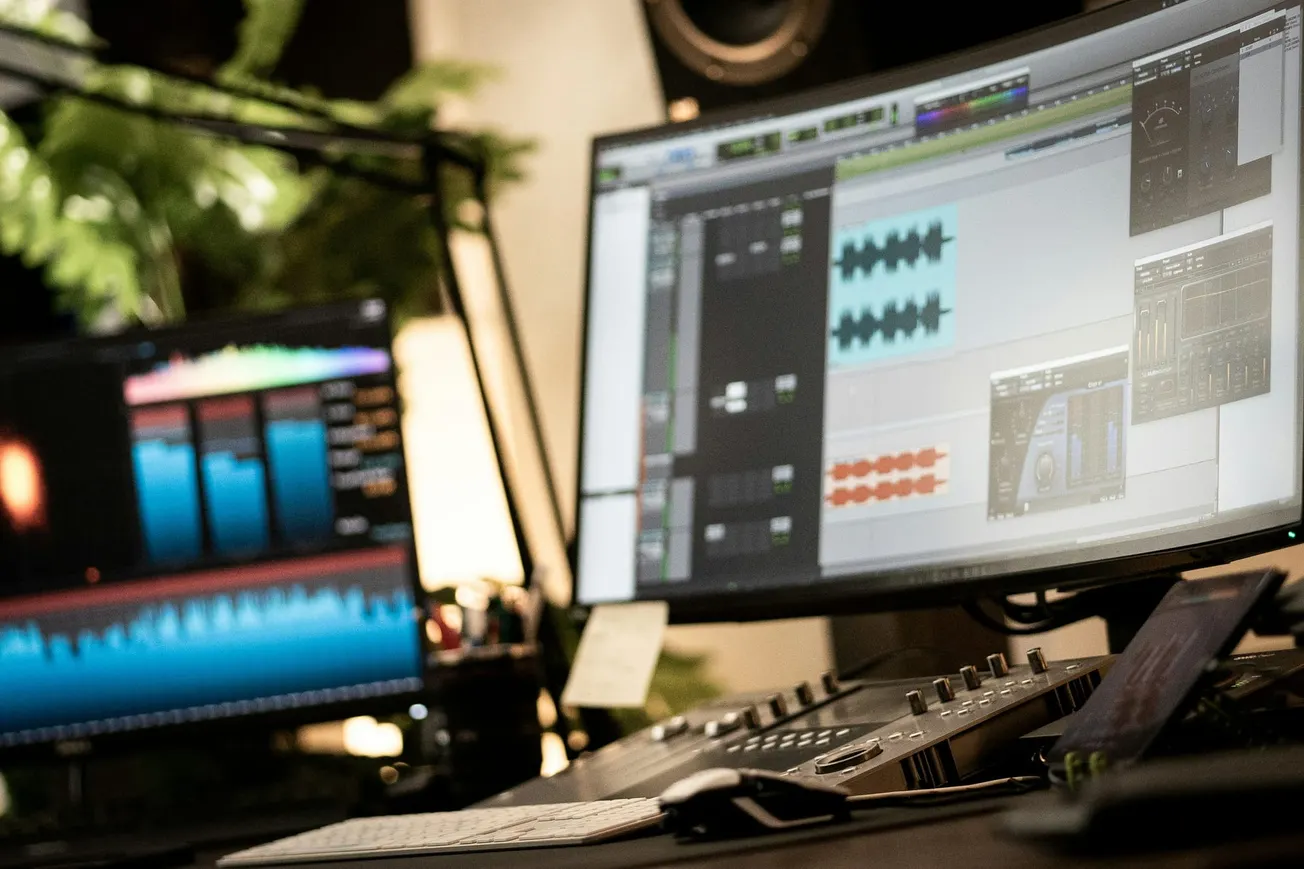 A music production studio with two monitors displaying audio waveforms and editing software. A keyboard and mixer are in the foreground; the setting feels technical and focused.