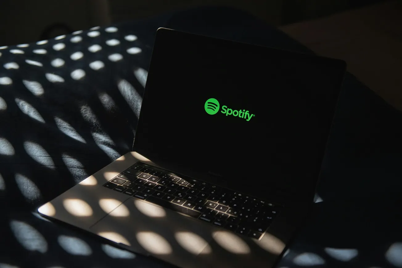 A laptop displaying the Spotify logo sits on a bed, with soft, dappled light patterns from a window creating a serene and cozy atmosphere.