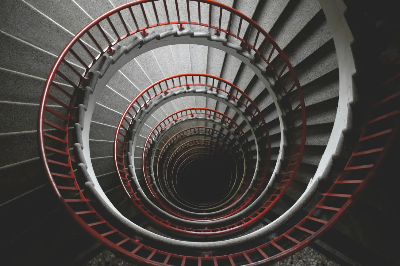 Aerial view of a spiral staircase with red railings and gray steps, descending into darkness, conveying depth, symmetry, and an intriguing visual pattern.