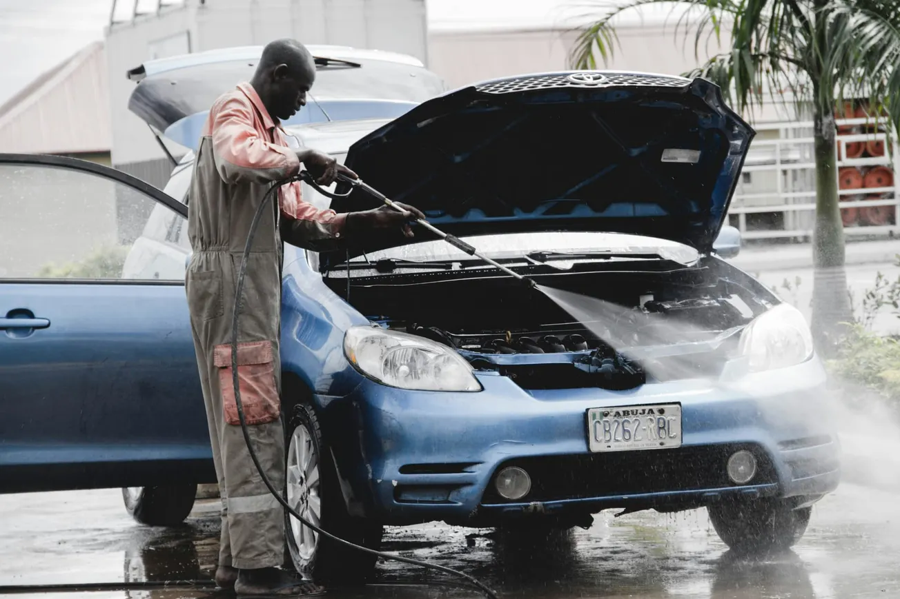 A person in overalls uses a pressure washer to clean the engine of a blue car with an open hood, set in an outdoor car wash area with palm trees.