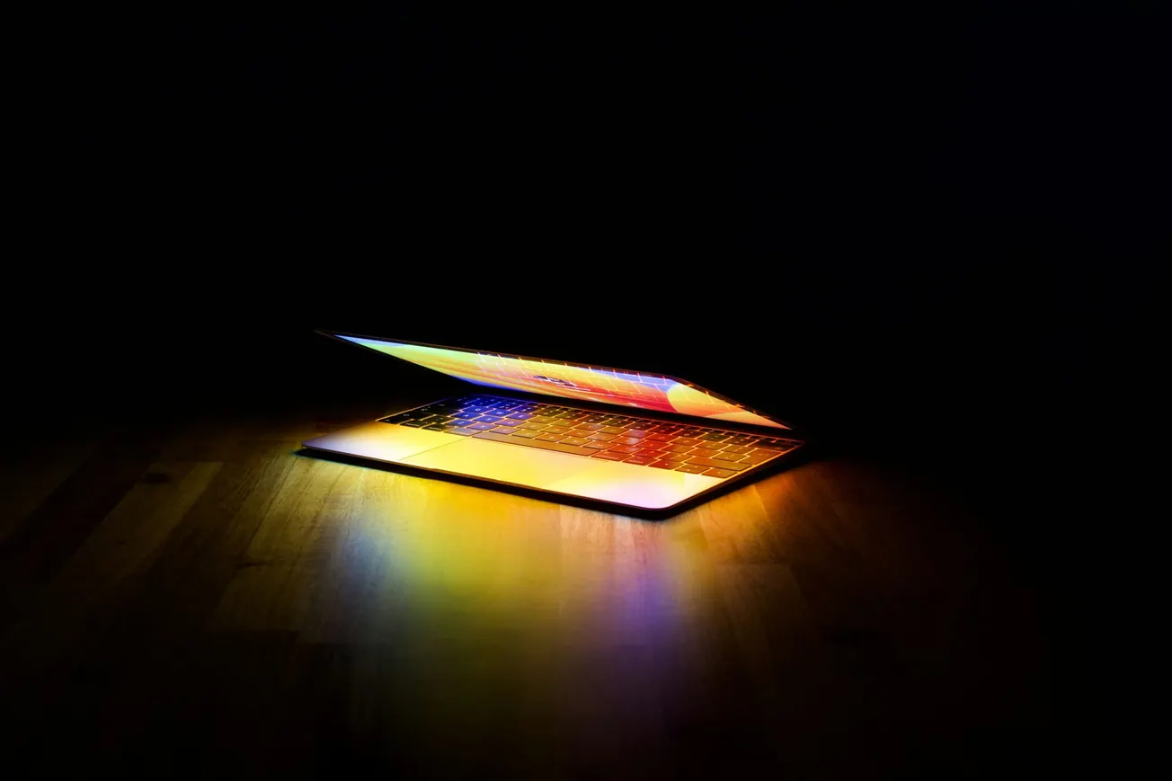 Semi-open laptop with a glowing screen casting vibrant colors on a wooden surface, set in a dark room, creating a mysterious ambiance.