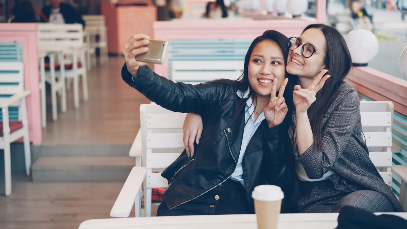Two friends smiling and taking a selfie at a cafe; one wears a black jacket, the other glasses. Bright, cheerful atmosphere with pastel decor.