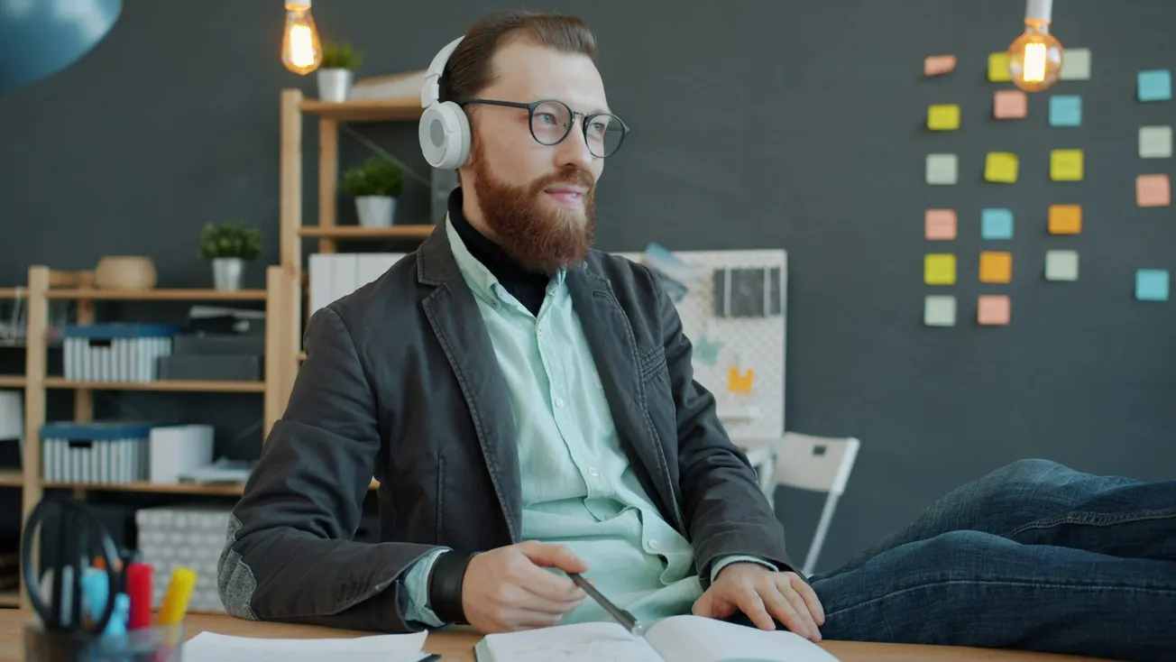 Man with headphones and glasses sits at a desk with a notebook, looking thoughtful. Colorful sticky notes are on the wall behind, creating a creative office vibe.