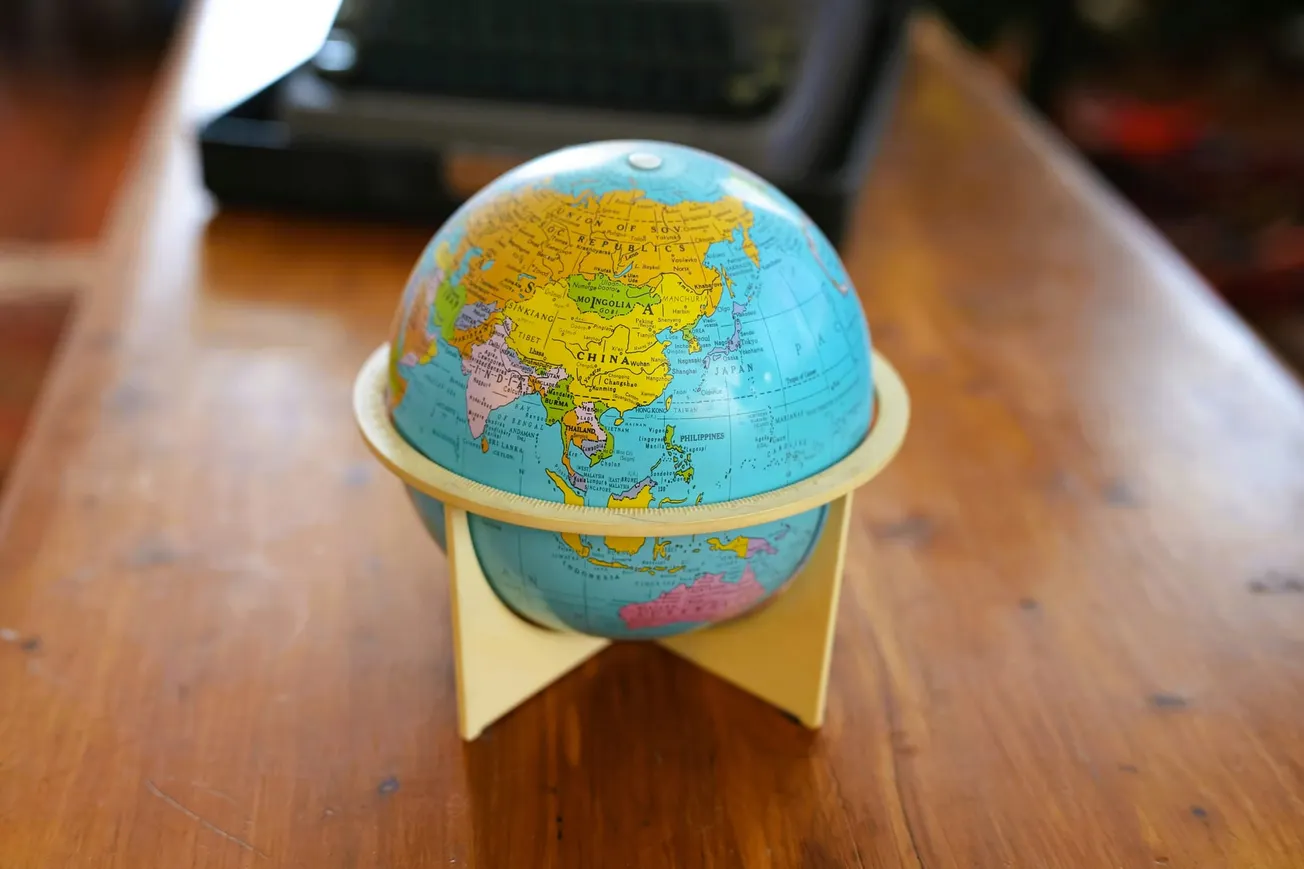 A small, colorful globe showing Asia and Australia sits on a wooden table, casting a soft shadow. A blurred laptop is visible in the background.