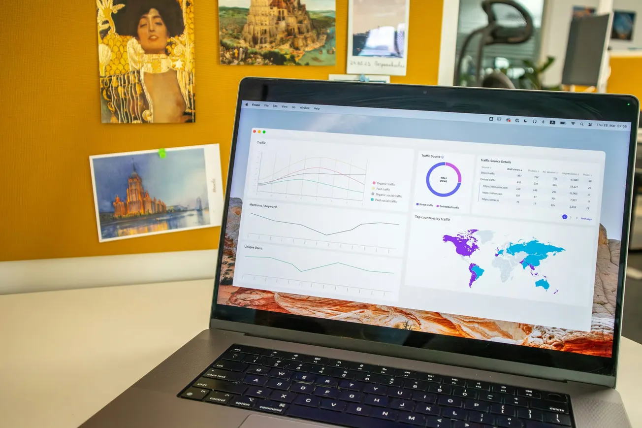 A laptop displaying analytics graphs and a world map sits on a desk. Background features an art-covered corkboard, creating a creative office vibe.