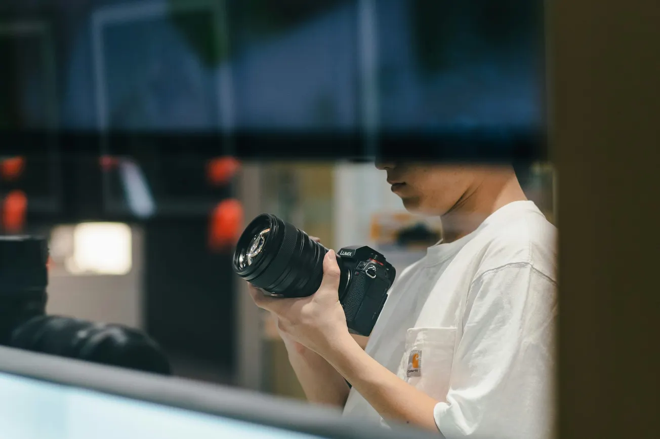 A person in a white shirt is holding a camera, partially obscured by a reflection. The image conveys focus and curiosity in a dimly lit room.
