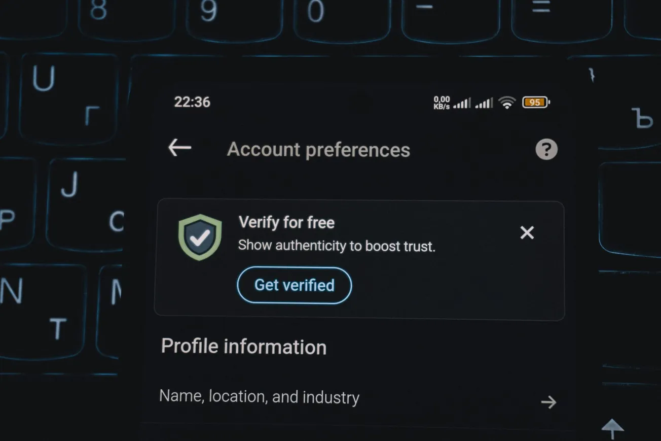 Close-up of a smartphone screen displaying "Account preferences" with an option to "Get verified" for free, overlaying a dimly lit keyboard.