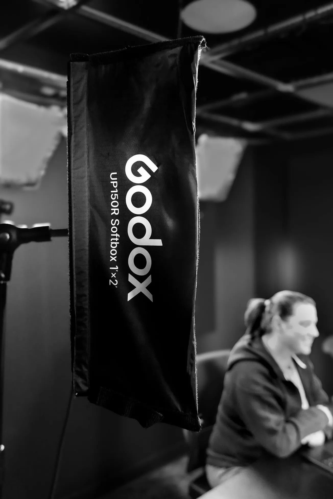 Black and white photo shows a Godox softbox in focus on the left, with the blurred figure of a person seated at a table in a dimly lit room.