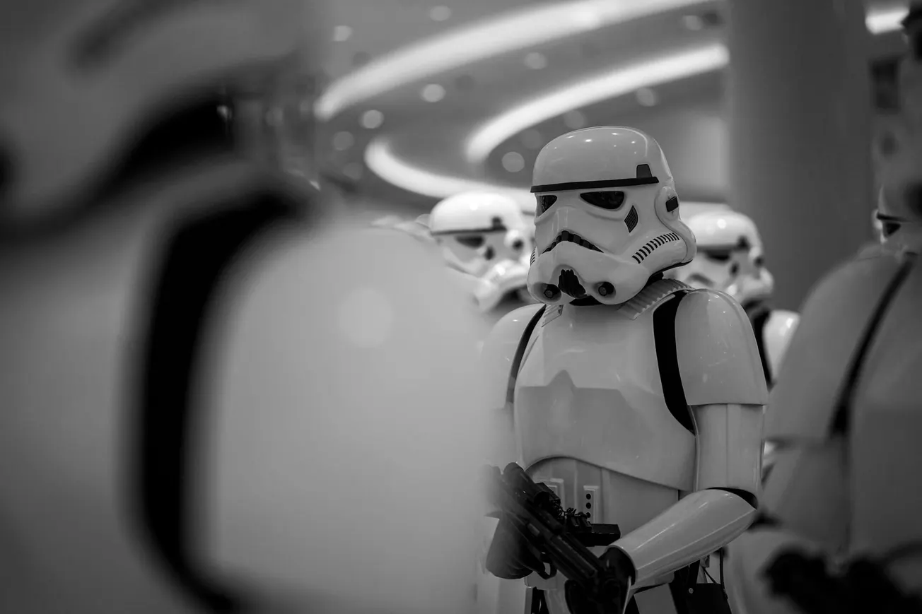 A group of Stormtroopers, fully armored in white, stands indoors under bright circular lights. The image is in black and white, creating a serious tone.