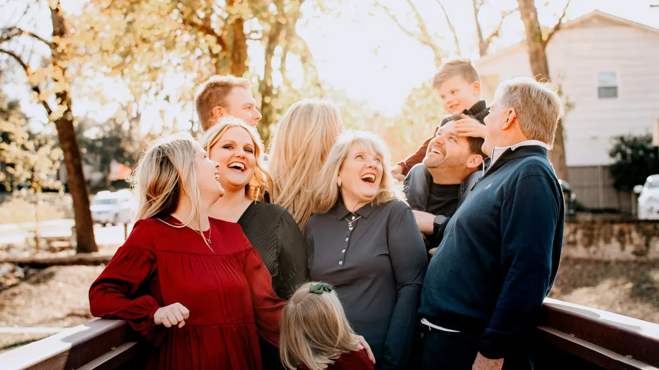 A joyful family gathers on a bridge in autumn, sharing laughter. Adults and children look happy, surrounded by fall foliage under a bright sky.