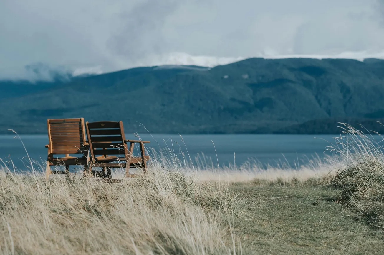 Wooden chairs sit on a grassy hill overlooking a serene lake and mountains under a cloudy sky, conveying tranquility and solitude.
