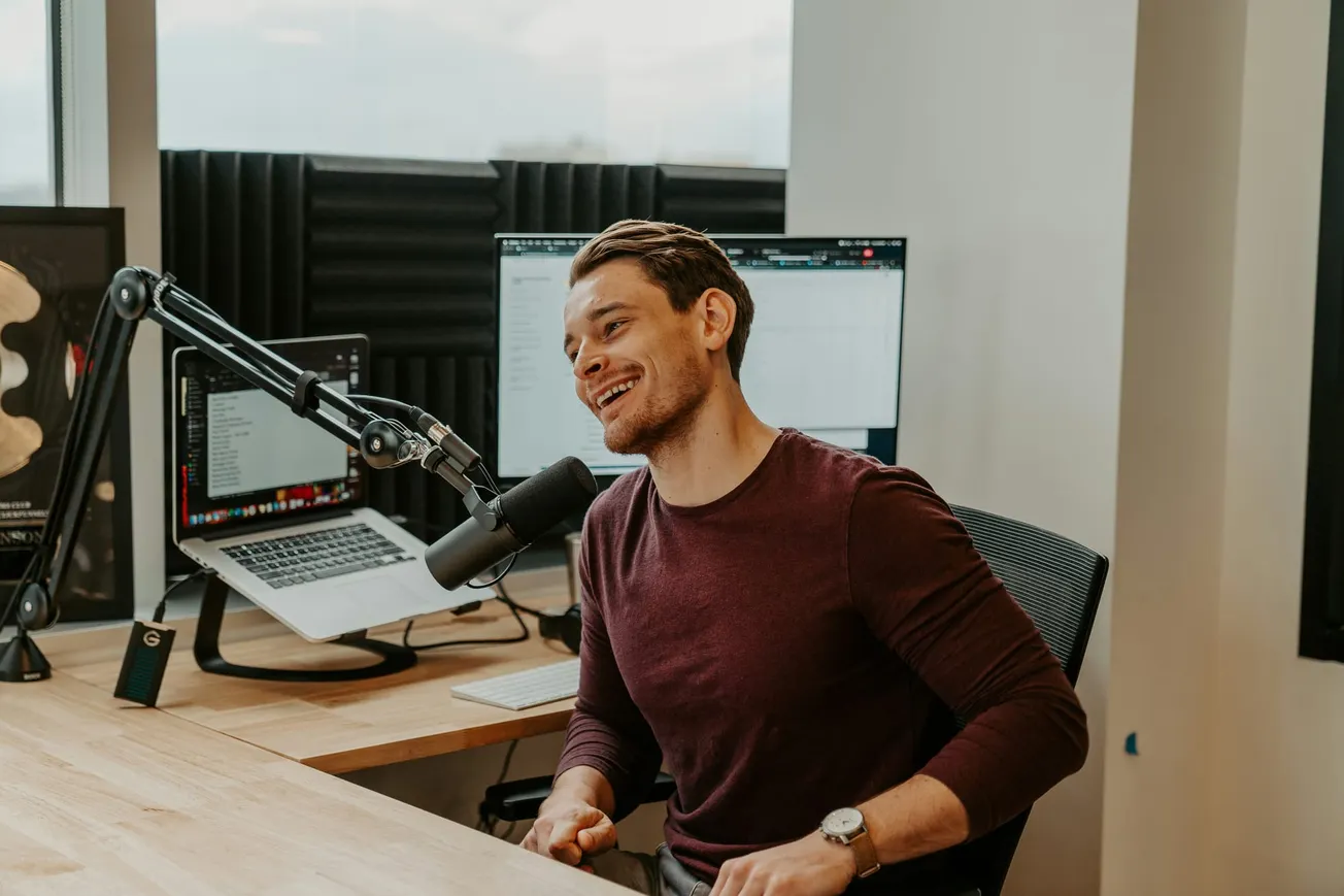 Man in a burgundy shirt sits at a desk with a microphone, smiling warmly. A laptop and monitor are on the desk. The setting is a modern office.