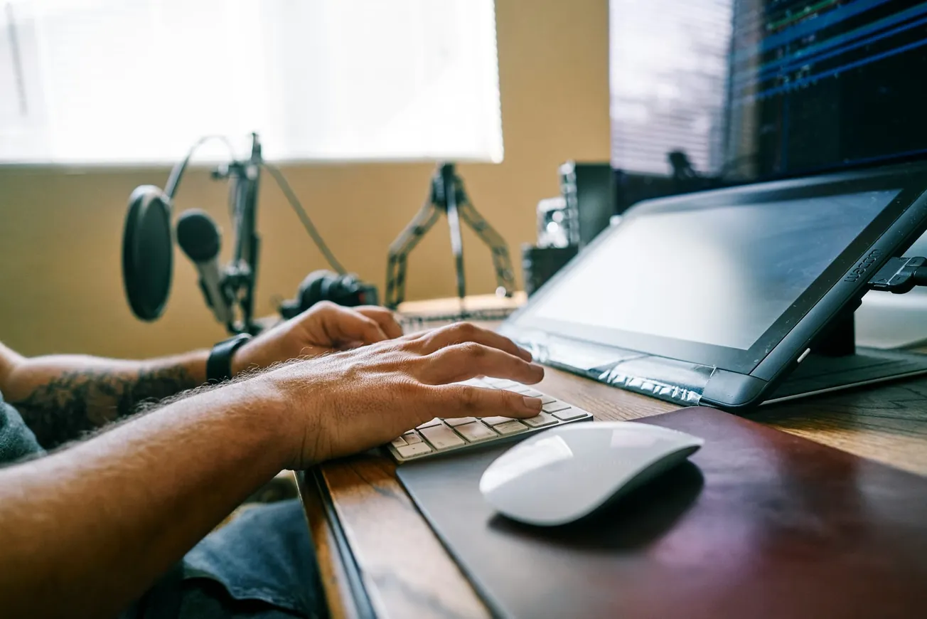 A person works at a wooden desk with a keyboard, mouse, and drawing tablet. A microphone and camera are visible in the background, suggesting a creative setup.