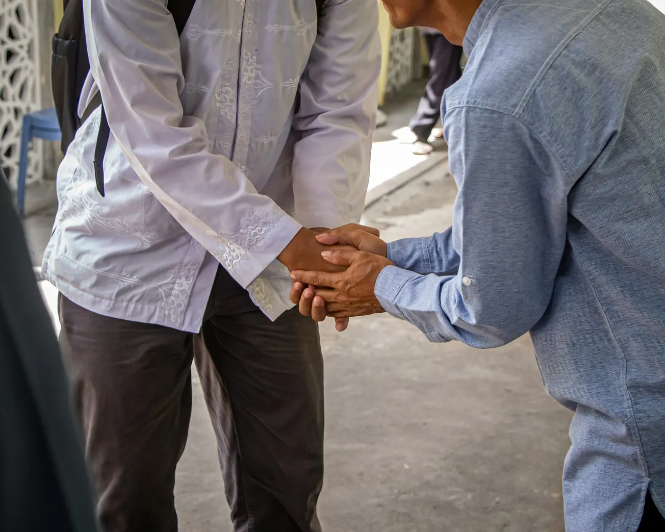 Two people exchange a warm handshake outdoors, one in a white shirt and the other in blue. The gesture conveys respect and friendliness.