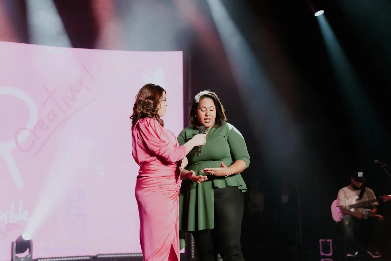 Two women stand on stage under spotlights. One in a pink dress hands a microphone to the other in green. A pink screen with text is in the background.