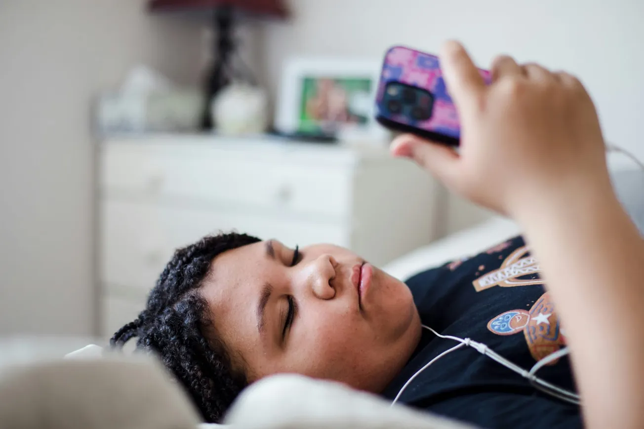 A person with braided hair lies on a bed, intently looking at a smartphone. Earbuds are in, suggesting they're listening to something. A bedside table is visible.