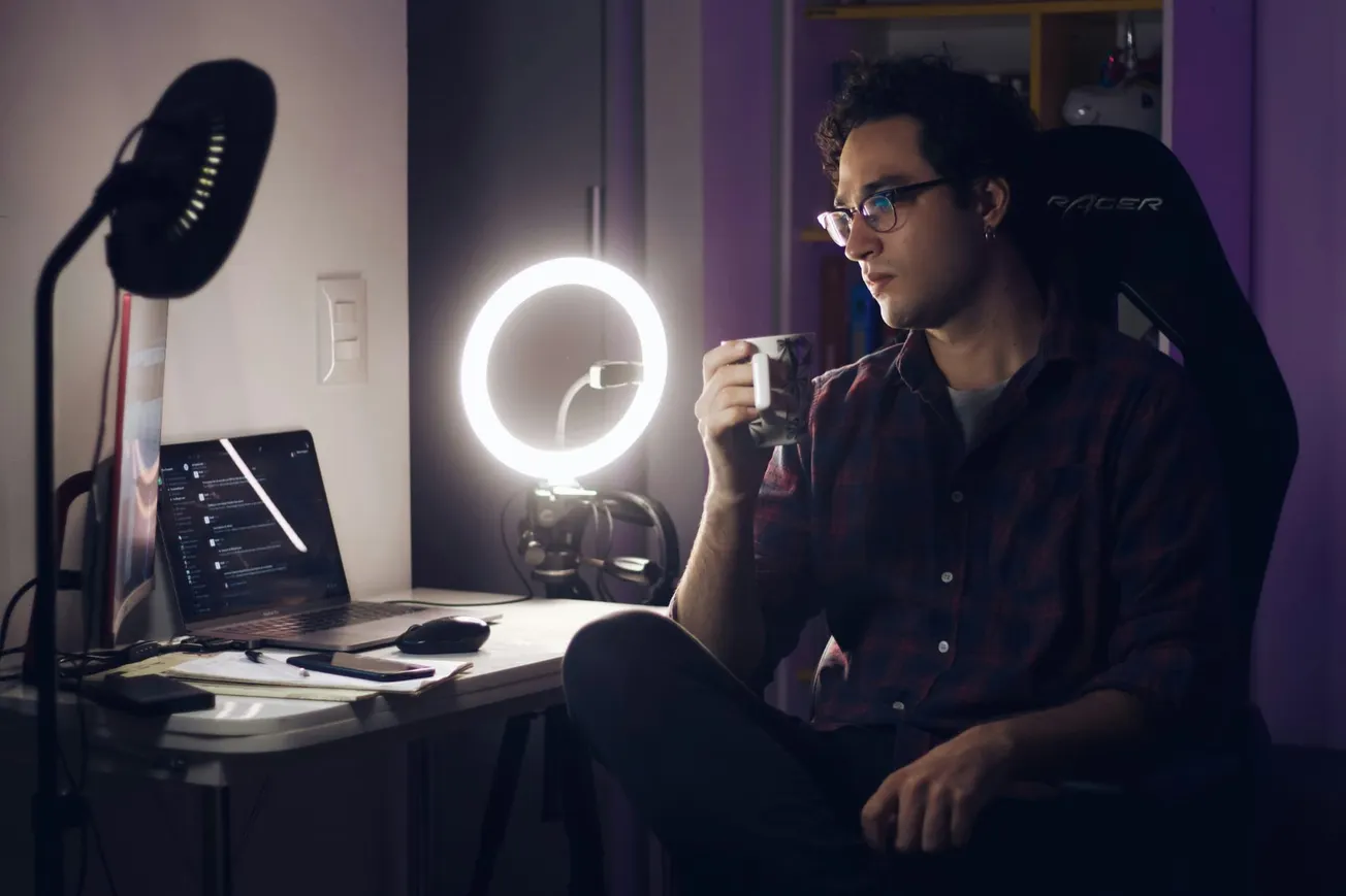 A person sits at a desk in a dimly lit room, drinking from a mug. A bright ring light and laptop illuminate the space, creating a cozy, focused atmosphere.