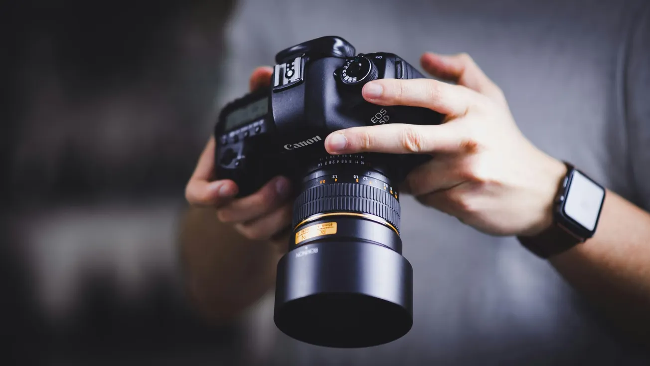 Close-up of hands holding a DSLR camera with a large lens. The focus is on the camera, conveying professionalism and attention to detail.