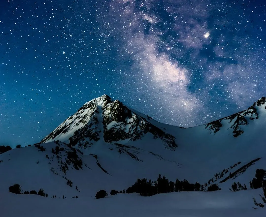 An astrophotographer operates a camera on a star tracker tripod, capturing a vibrant Milky Way arching over rugged mountains and distant city lights under a dark, clear night sky.