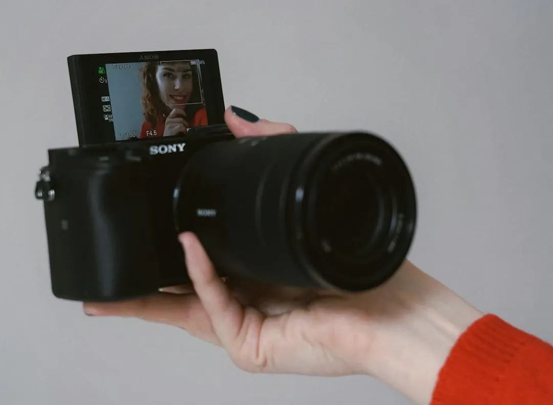 A person's hand holds a sleek silver compact camera, actively recording video footage in a bright, modern studio environment.