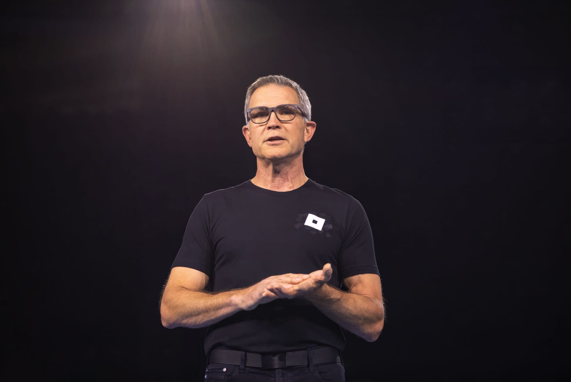 Roblox CEO David Baszucki standing and speaking against a black backdrop. He's holding his hands as if rubbing them together.