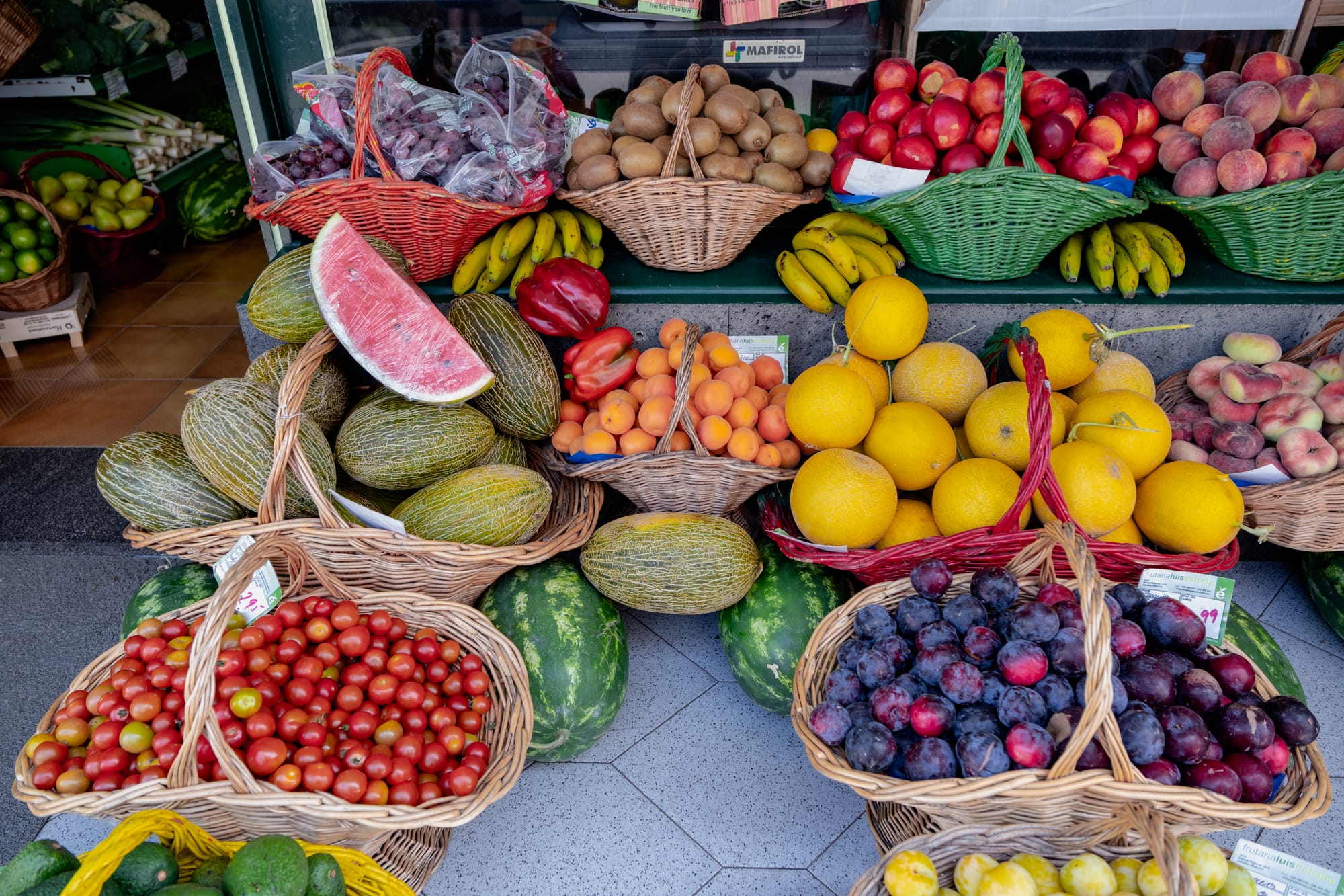Wicker baskets filled with plums, tomatoes, melons, and peaches outside Frutaria Luís Estrela in Furnas