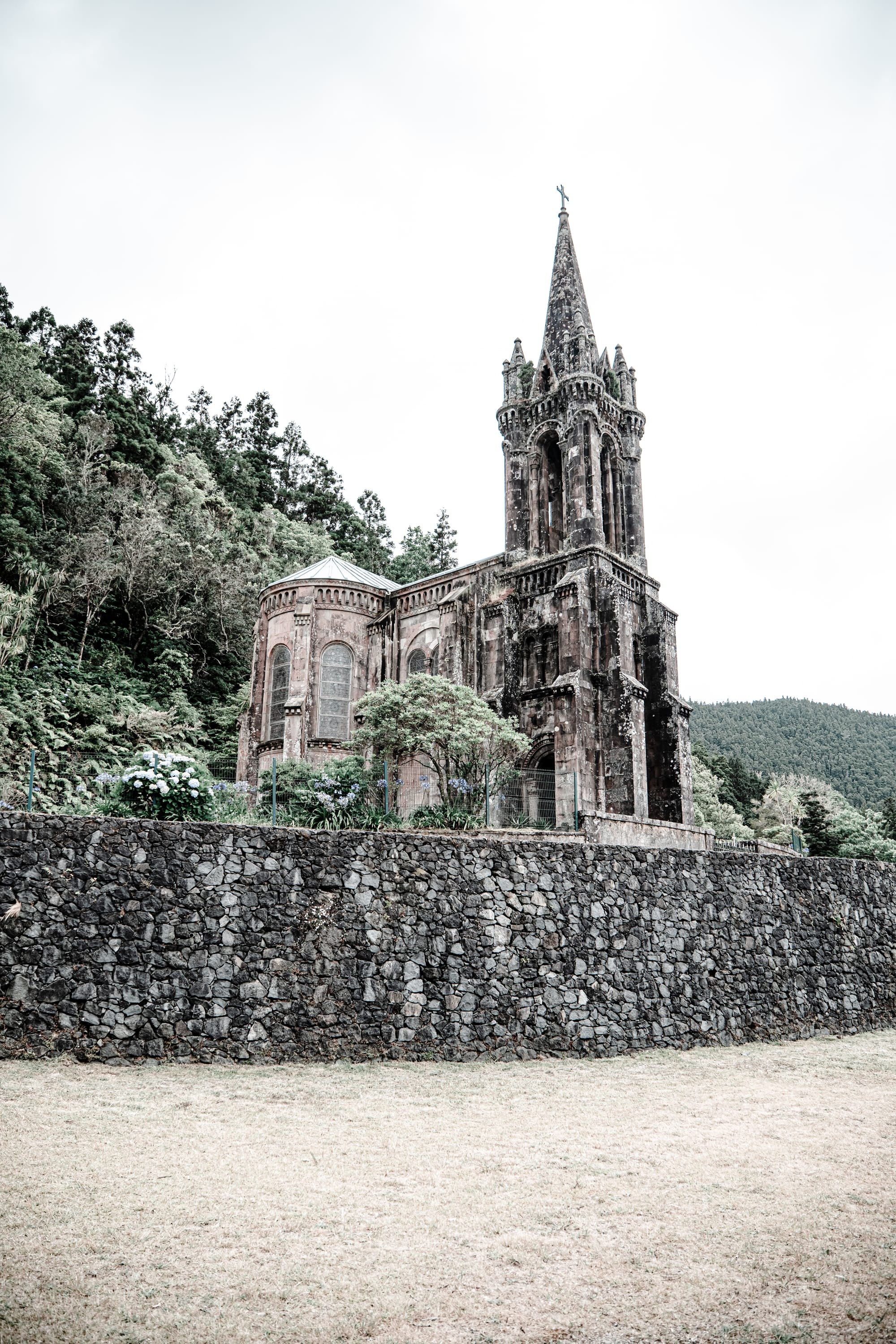 A tall stone chapel with a pointed spire and arched windows sits above a stone wall, backed by forested hills near Lagoa das Furnas