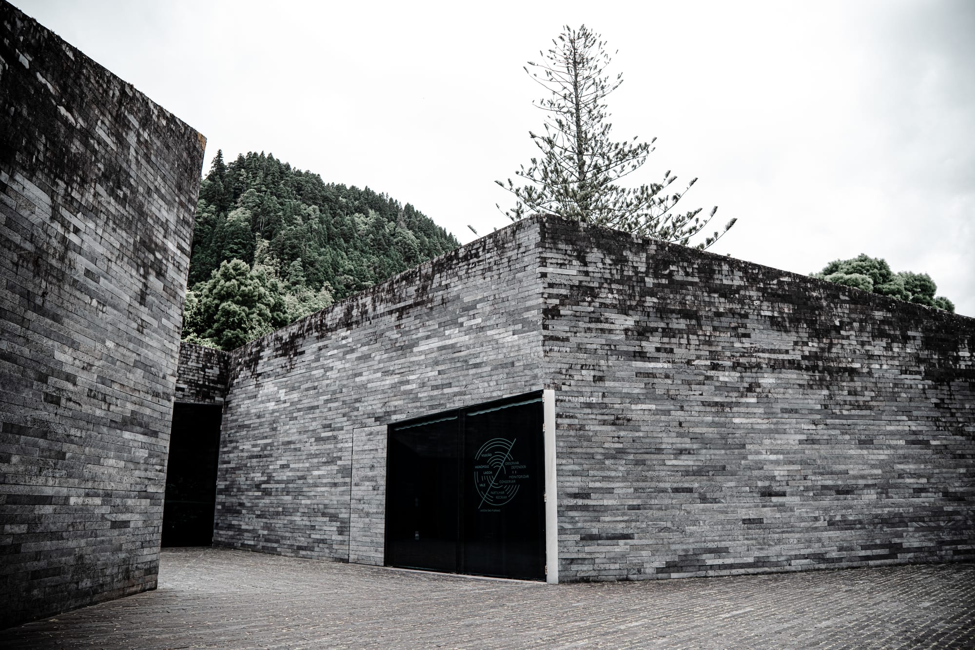 Modern grey-brick building of the Furnas Monitoring and Research Center, set against forested hills and cloudy sky