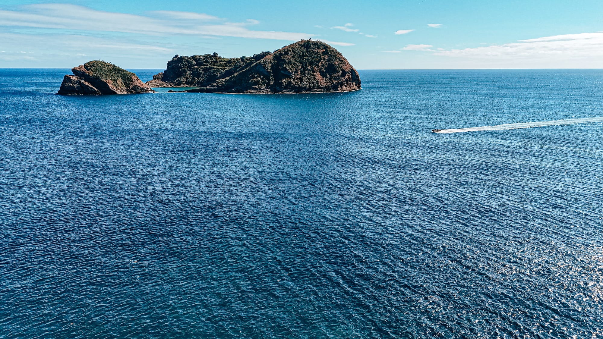 A wide aerial view of Ilhéu de Vila Franca do Campo with a small speedboat cutting across the ocean in the foreground
