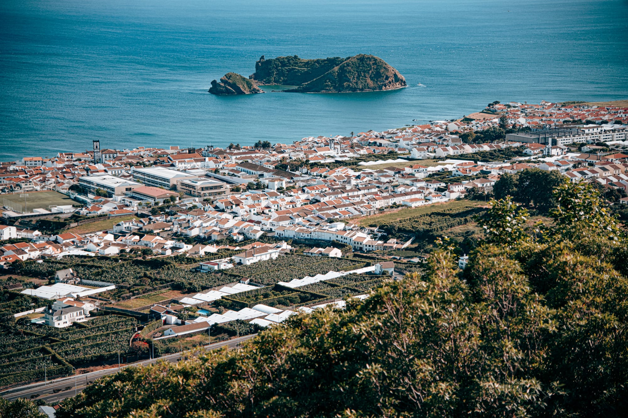A wide elevated view showing the town of Vila Franca do Campo in the foreground with its white houses and red roofs, and Ilhéu de Vila Franca do Campo floating in the Atlantic beyond