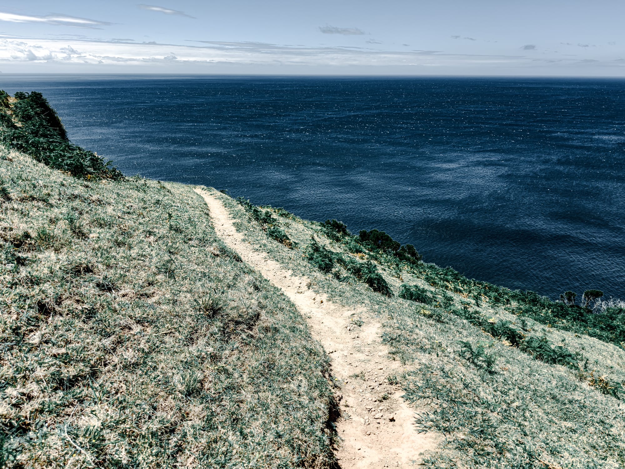 Dirt path winding through dry grass on a coastal cliffside, with the vast Atlantic Ocean stretching out beneath a partly cloudy sky