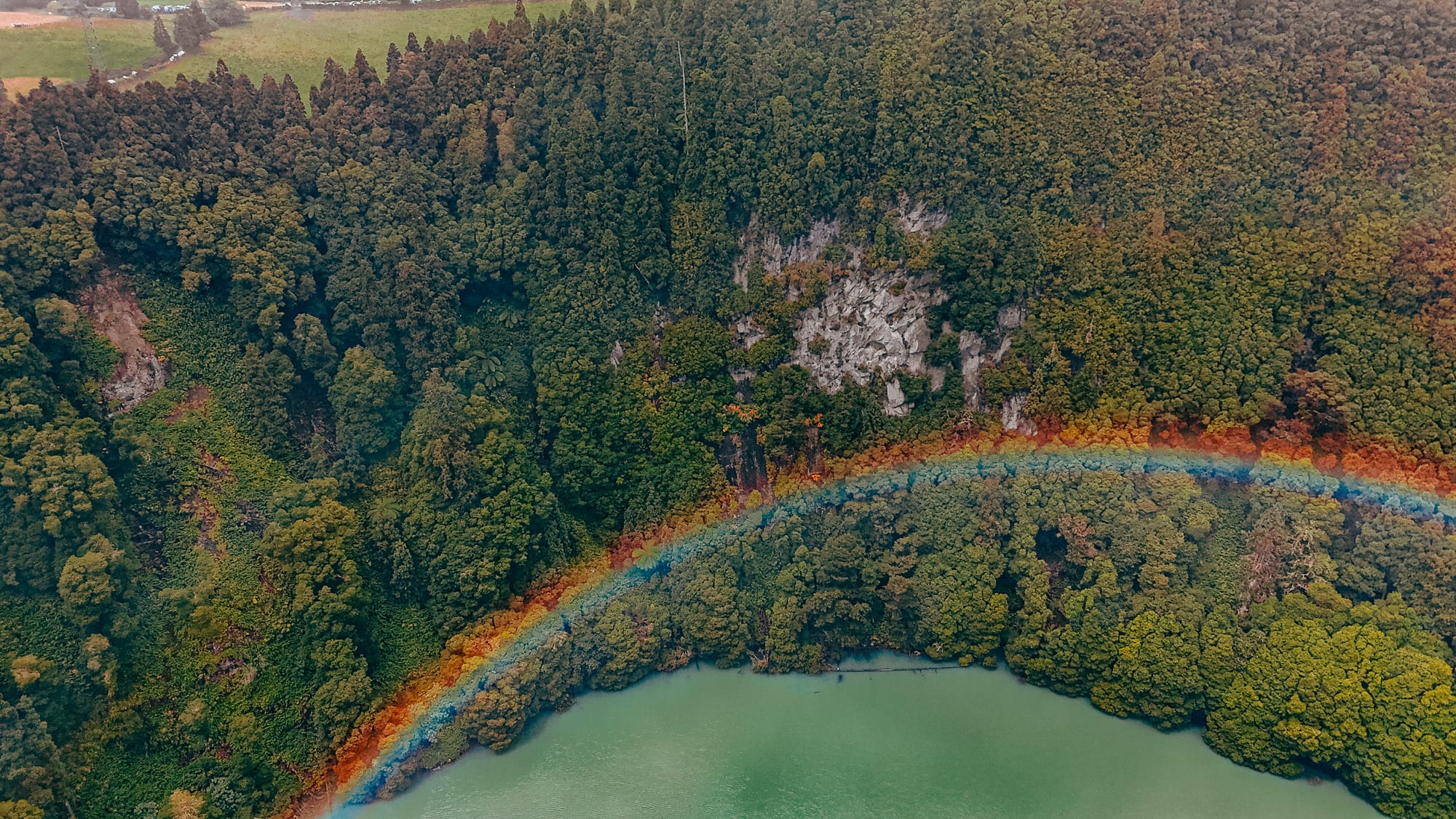 Aerial view of a lagoa on São Miguel, Azores, with a vivid rainbow stretching across dense green forest along the lake’s edge
