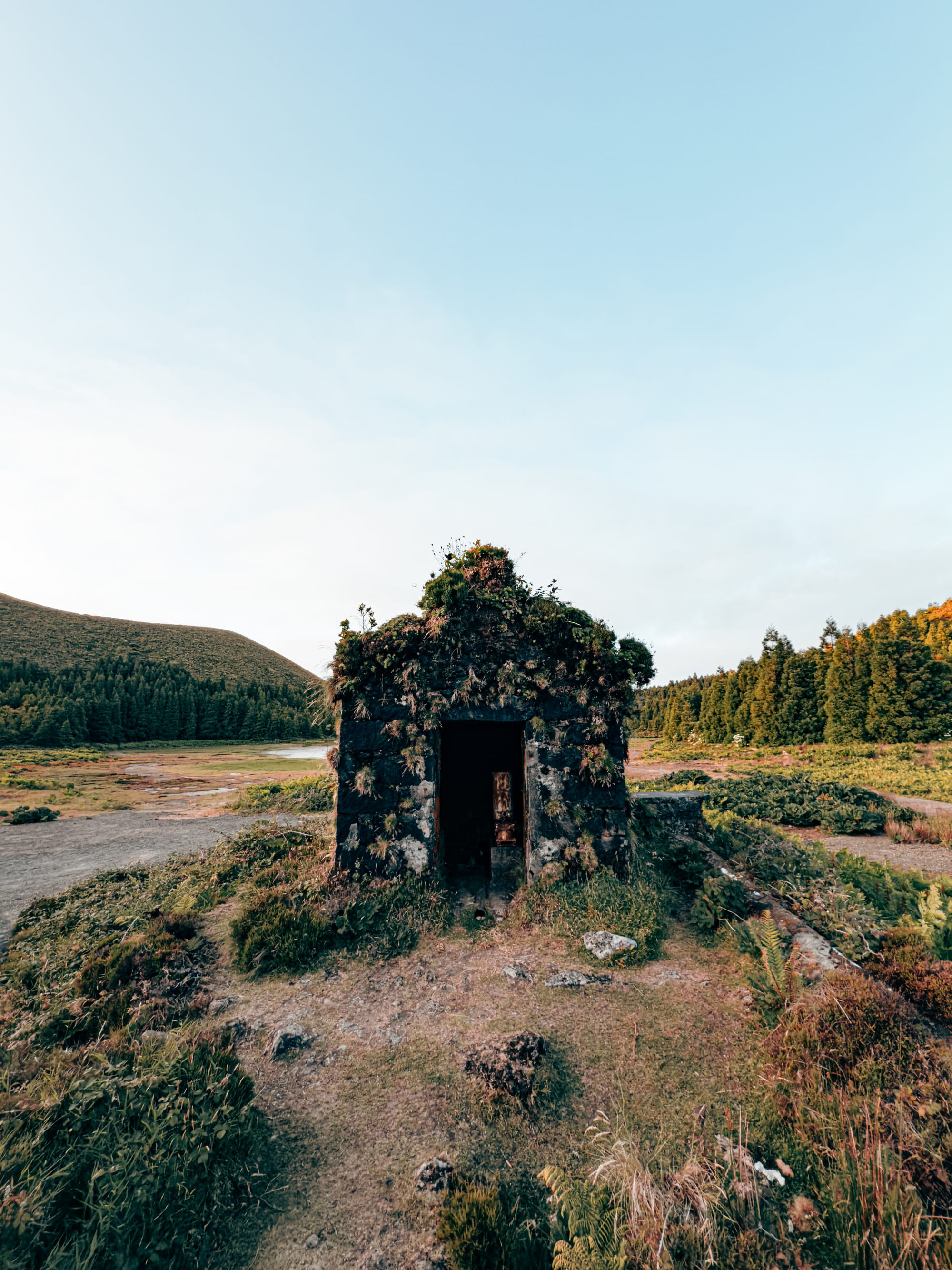 Small stone shelter with moss and plants growing on its roof and walls, situated in a volcanic landscape near a crater lake surrounded by forest