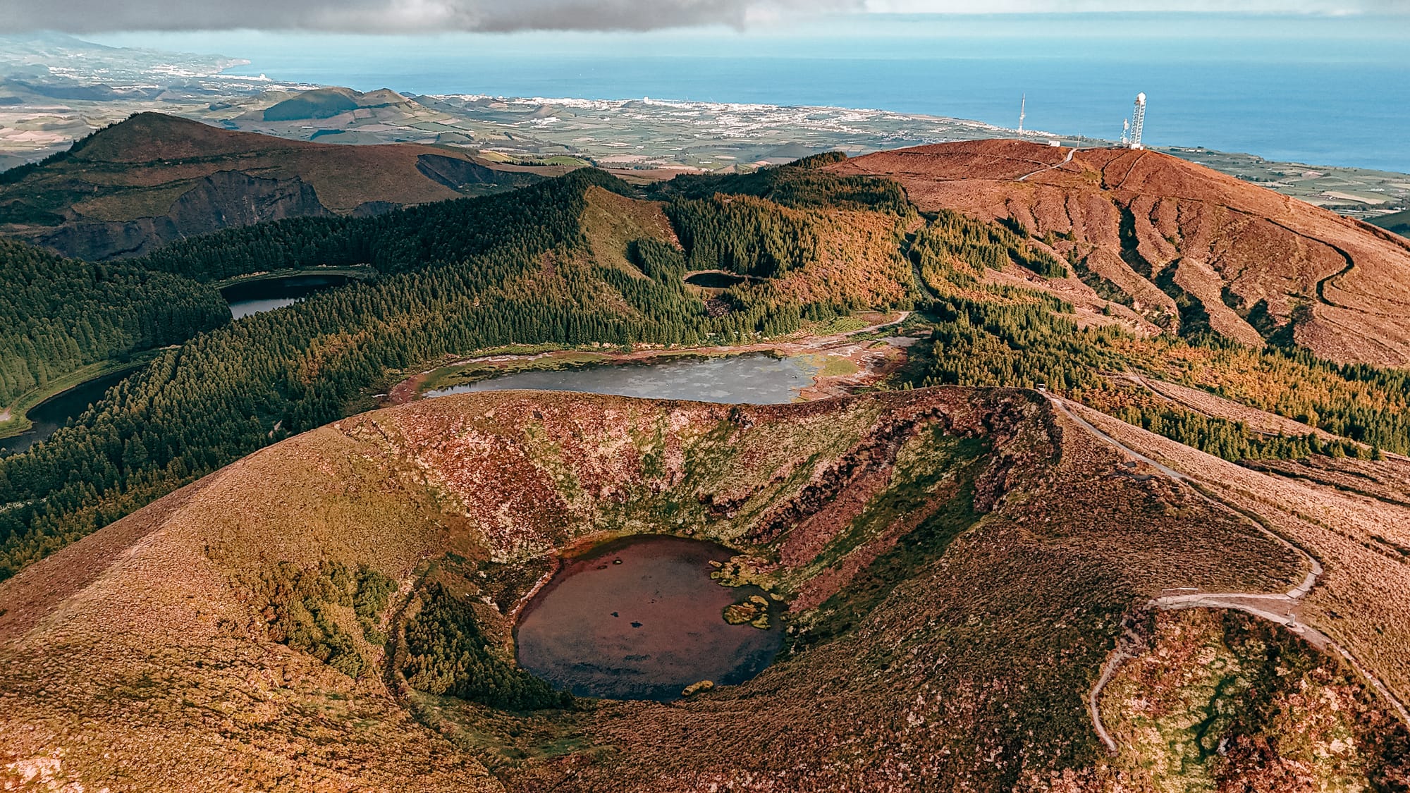 Aerial view of a volcanic region with three distinct crater lakes surrounded by ridgelines, pine forests, and winding trails, with the coastline and town visible in the distance under a pale sky