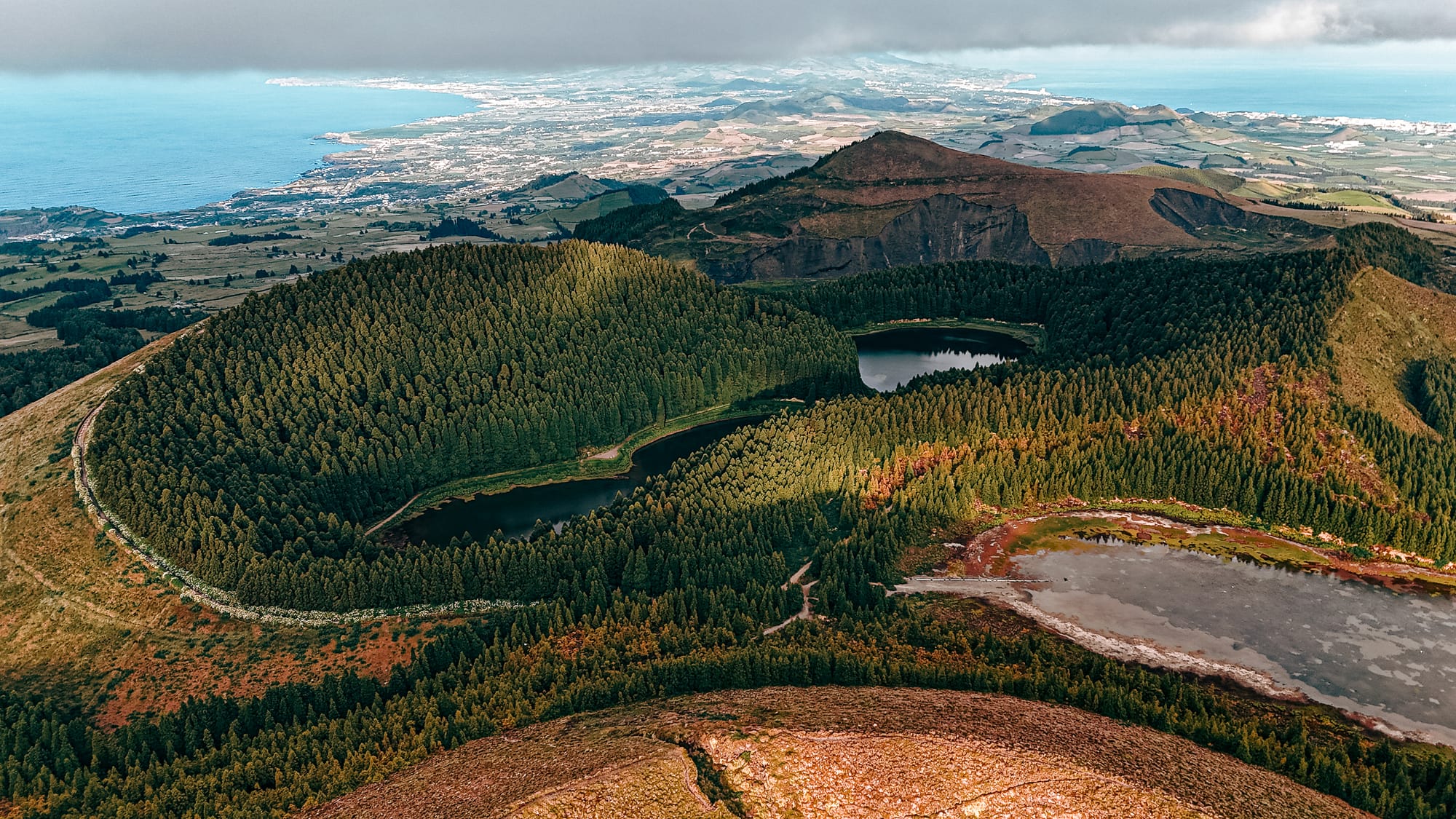 Aerial view of three crater lakes surrounded by lush conifer forest, with volcanic ridges, a coastal town, and the Atlantic Ocean visible in the distance