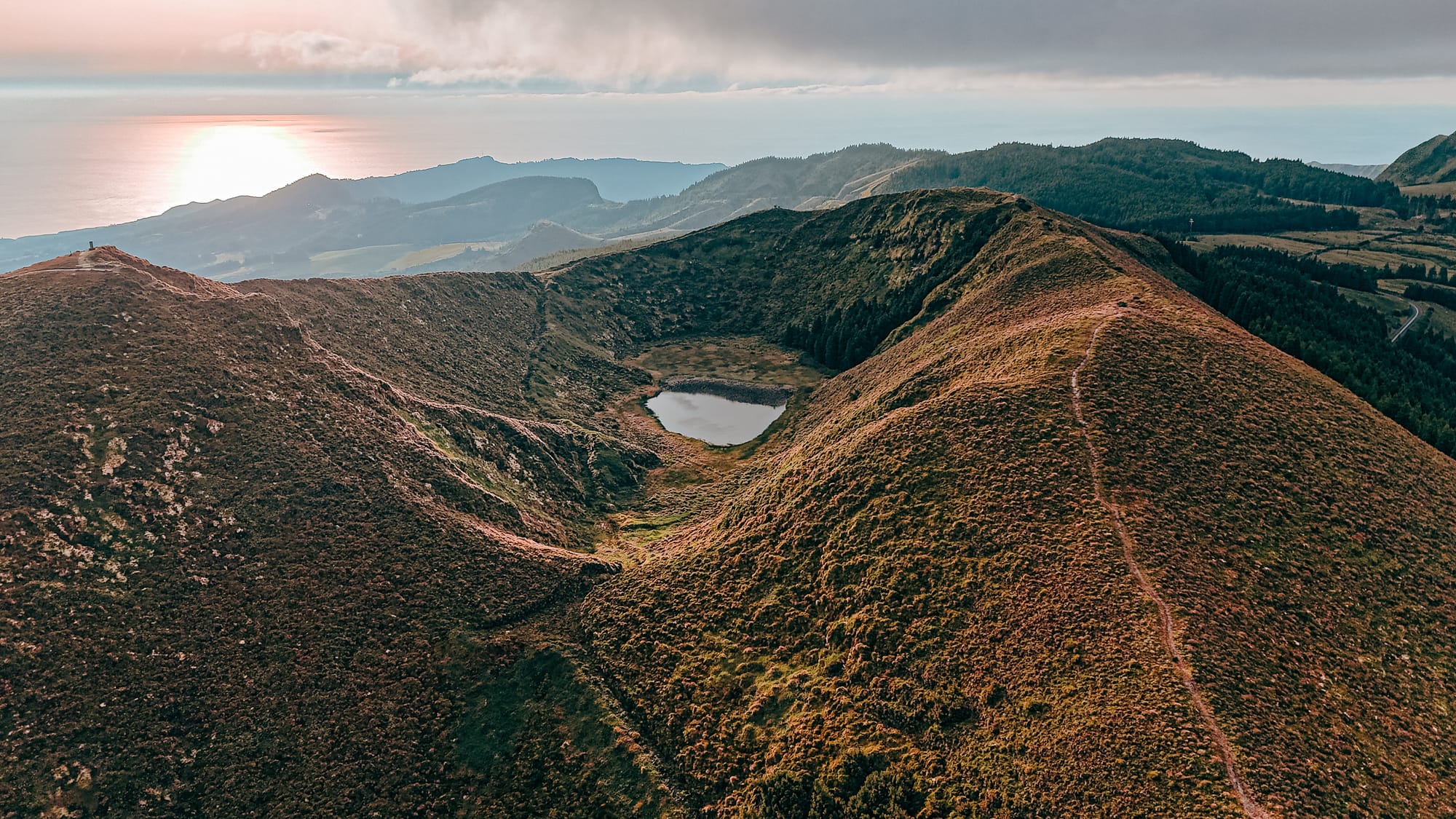 Aerial view of a crater lake encircled by rugged volcanic hills at sunset, with the Atlantic Ocean glowing in the distance