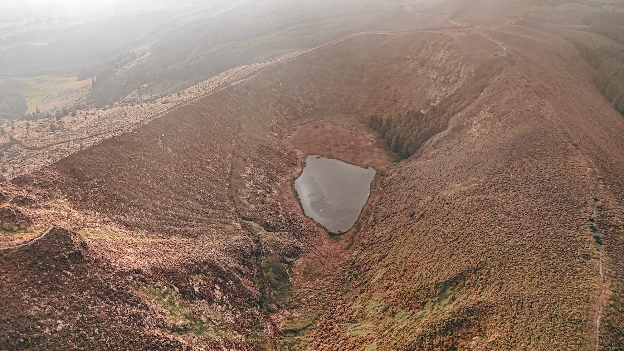 Aerial view of a heart-shaped crater lake nestled within a reddish-brown volcanic landscape, with soft morning light and a narrow trail winding along the ridge