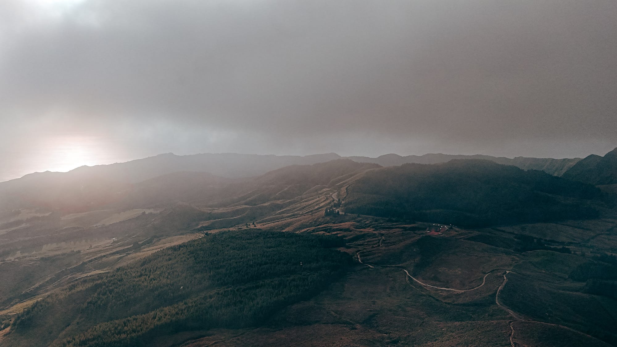 Distant view of a winding volcanic landscape under a glowing sunset, with soft clouds and atmospheric light spilling over forested ridgelines and open terrain